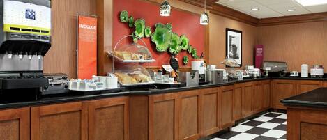 A hotel breakfast area with a coffee machine, a fruit display, and a menu board.