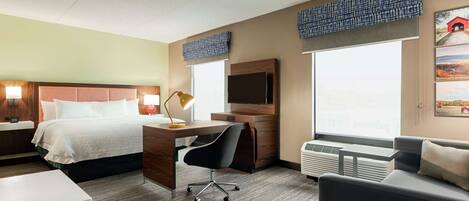 Una habitación de hotel con cama, escritorio, silla, televisor y ventanas amplias.