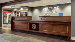 A reception desk with a "WELCOME THE CRAB STATE" sign, a Hilton Honors logo, and a view of a refrigerated beverage display in the background. - Hampton Inn & Suites Frederick-Fort Detrick (Frederick)