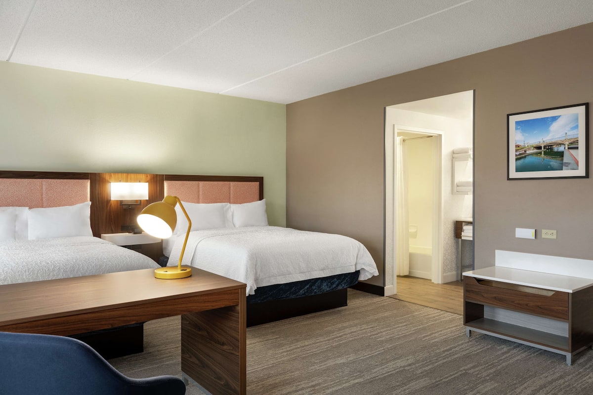 Ein Hotelzimmer mit zwei Betten, einem Schreibtisch mit Lampe, einem Nachttisch und einem durchgegangene Tür erkennbaren Badezimmer.