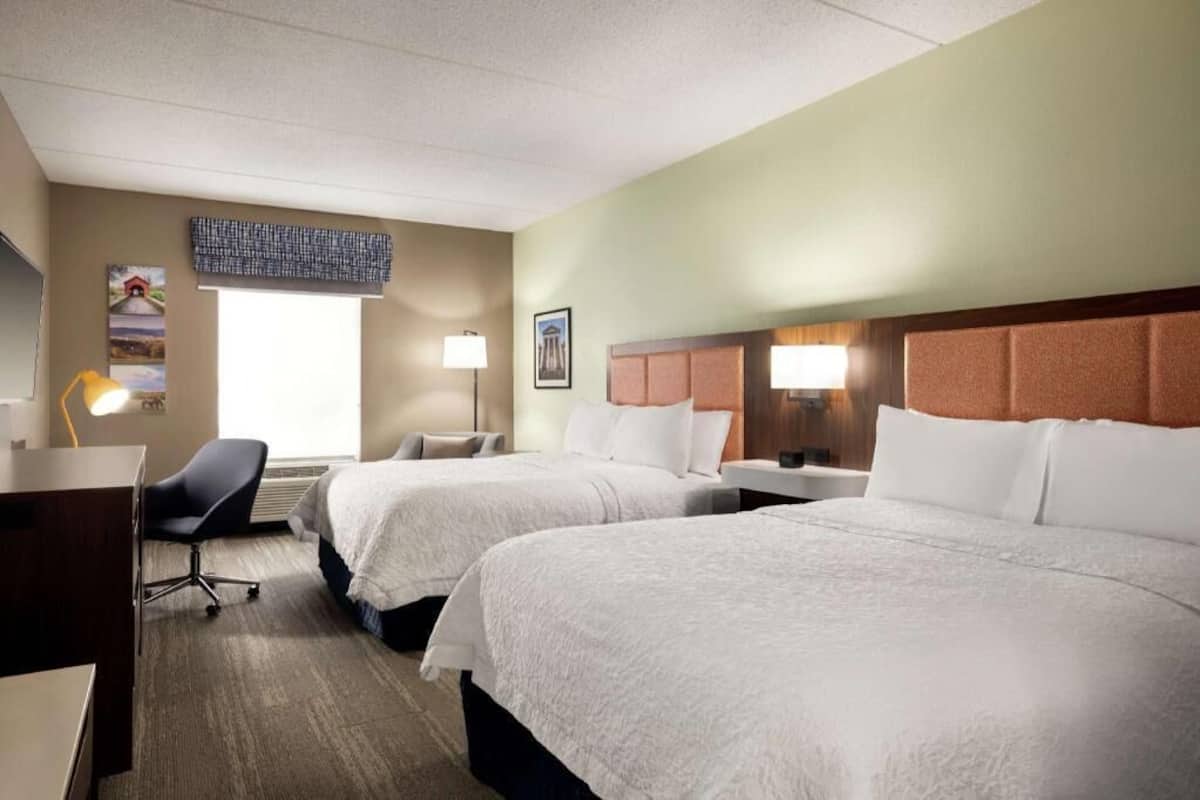 Ein Hotelzimmer mit zwei Betten, einem Schreibtisch, einem Stuhl und einem Fenster mit Jalousien.