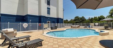 Una piscina con sillones de descanso y sombrillas, rodeada por una valla y un edificio con base azul.