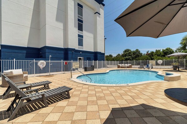 Une piscine entourée d’une clôture, agrémentée de chaises longues et de parasols, et bordée d’un bâtiment dont la base est bleue.