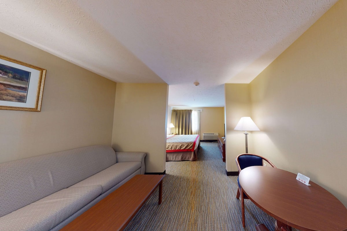 A hotel room with a sofa, a wooden table, a bed with a red bedspread, a lamp, and a framed picture on the wall.