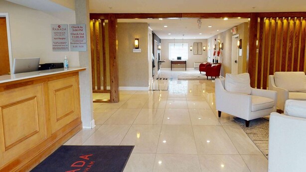 A hotel lobby with a reception desk, a check-in counter, a seating area with chairs, and a dining area in the background.