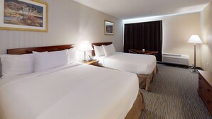 A hotel room with two beds, a nightstand with a lamp, a desk with a chair, a window with curtains, and a wall-mounted air conditioner.