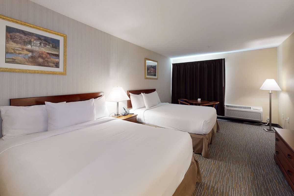 A hotel room with two beds, a nightstand with a lamp, a desk with a chair, a window with curtains, and a wall-mounted air conditioner.