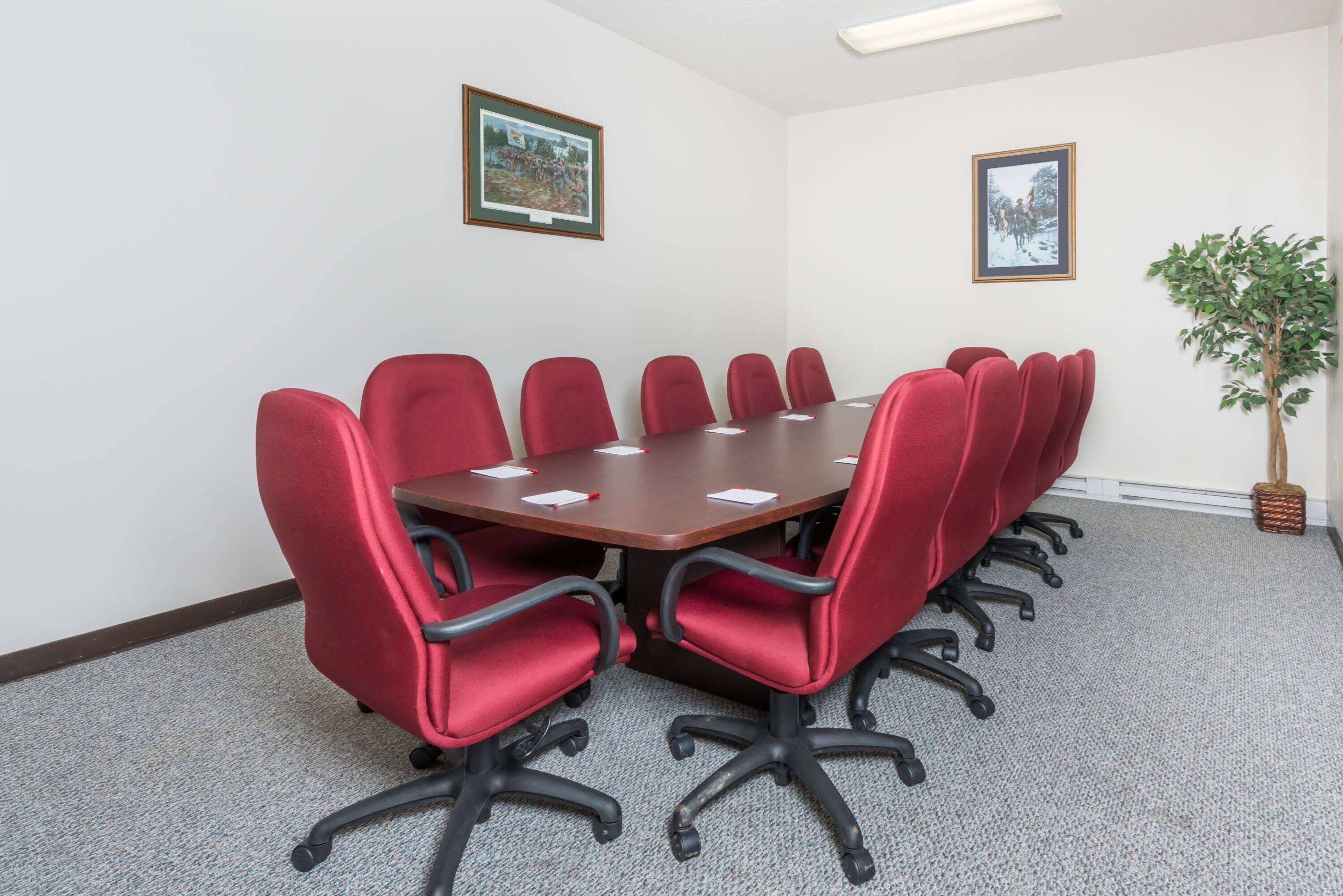 Une salle de conférence avec une table rectangulaire entourée de chaises rouges, des murs blancs et des tableaux encadrés.