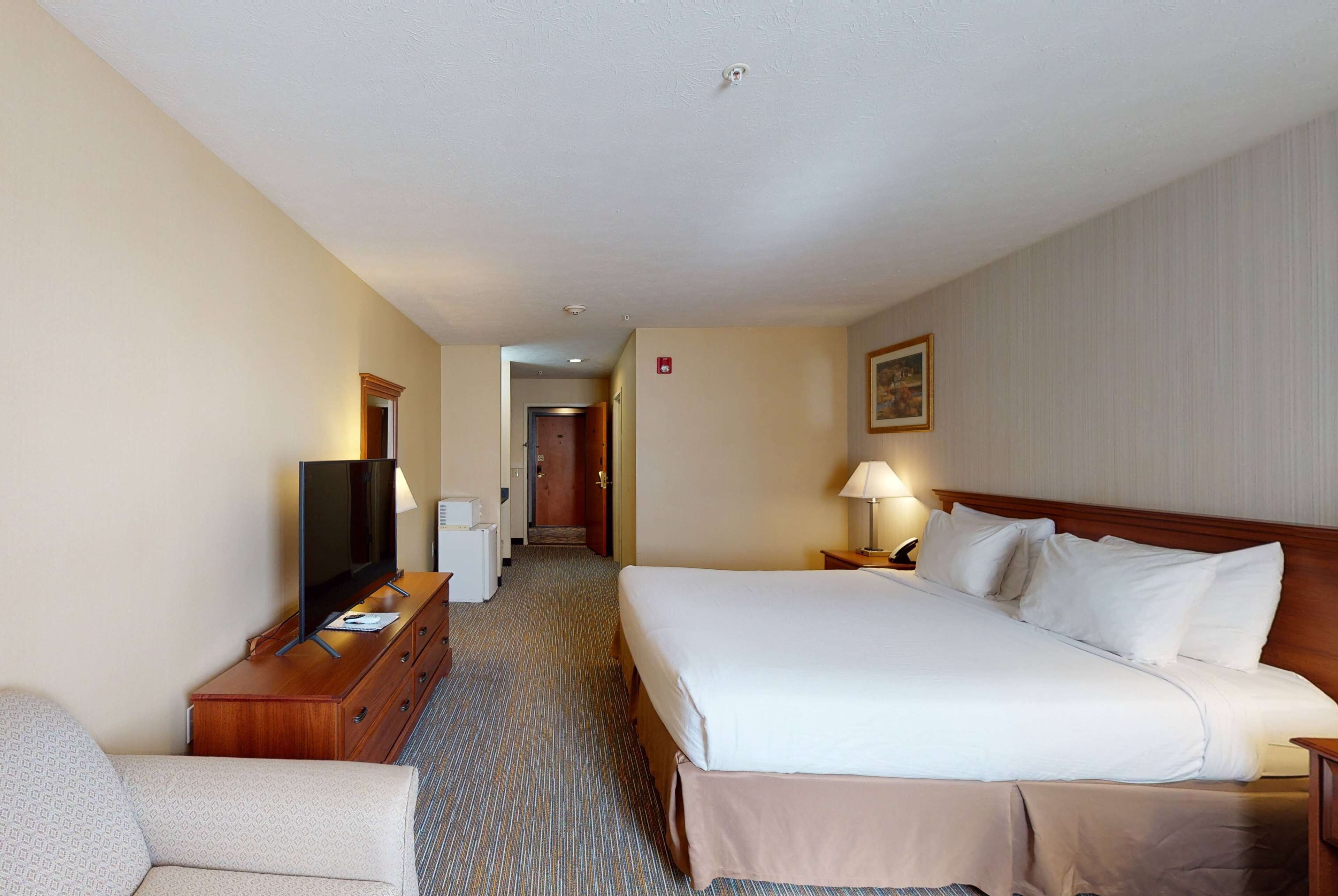 A hotel room with a large bed, a TV on a wooden dresser, a desk with a lamp, and a door leading to another room.