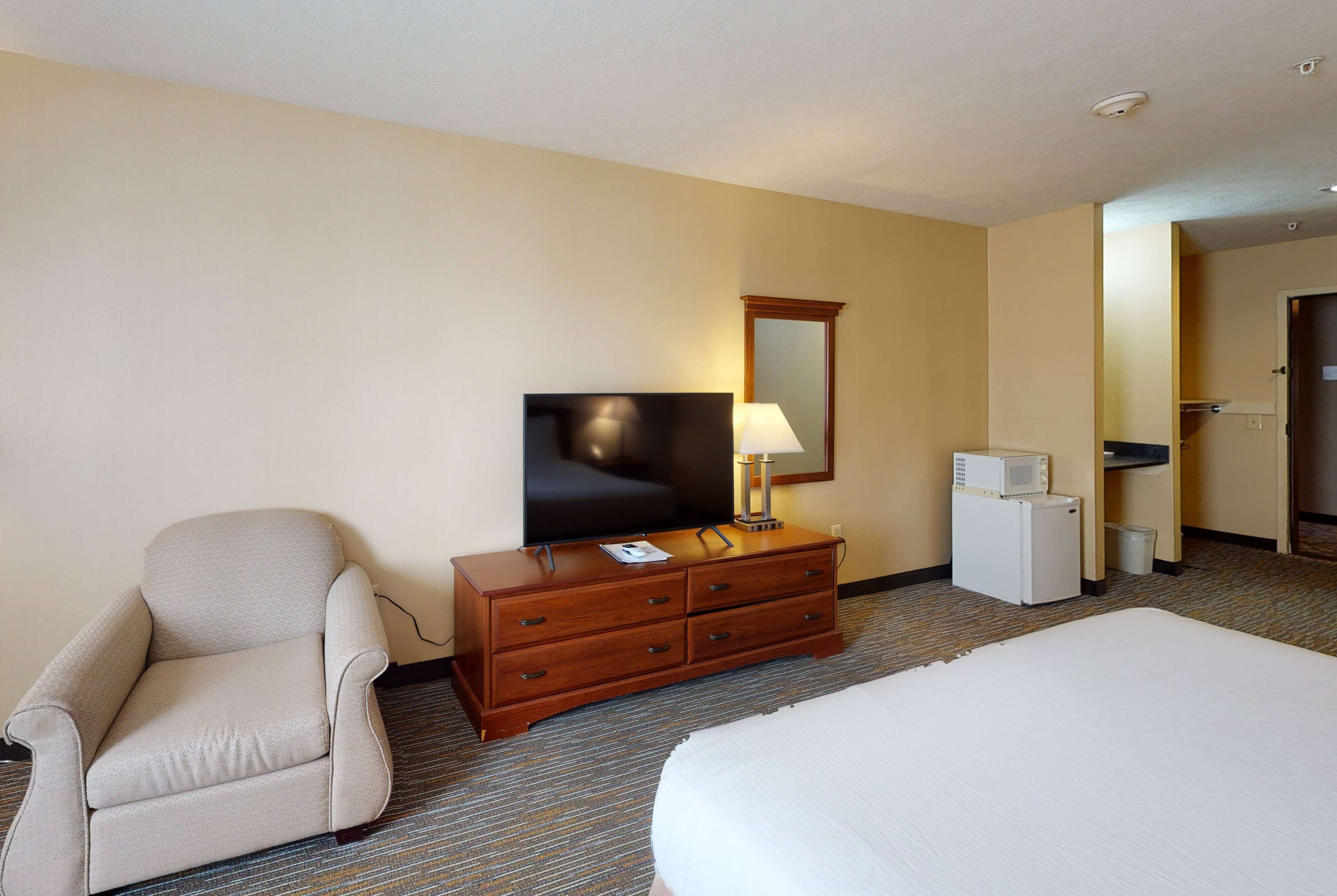 Habitación de hotel con cama, una silla, cómoda con televisor y lámpara, microondas y un refrigerador pequeño.