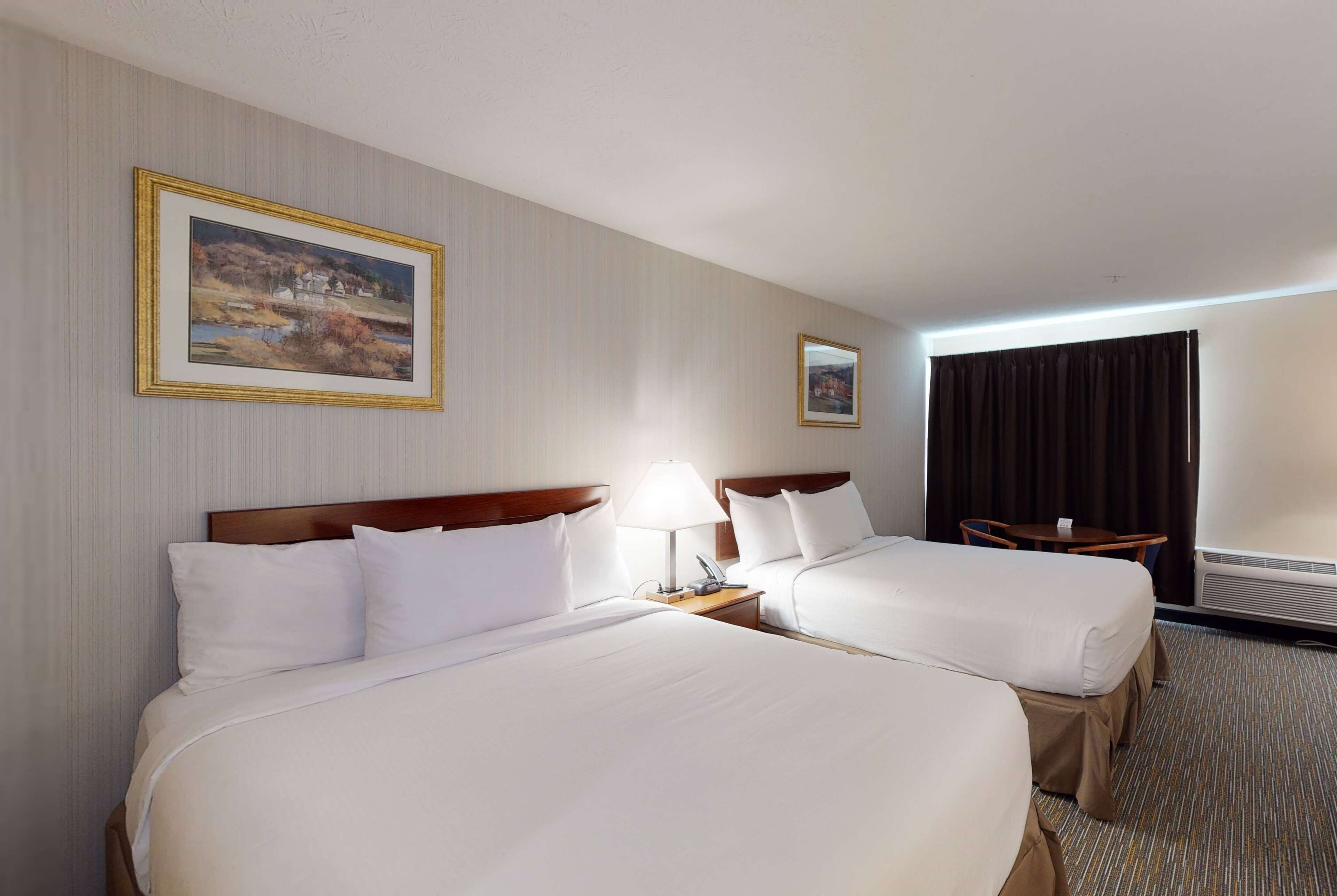 A hotel room with two beds, a nightstand with a lamp, a small table, and a painting on the wall.