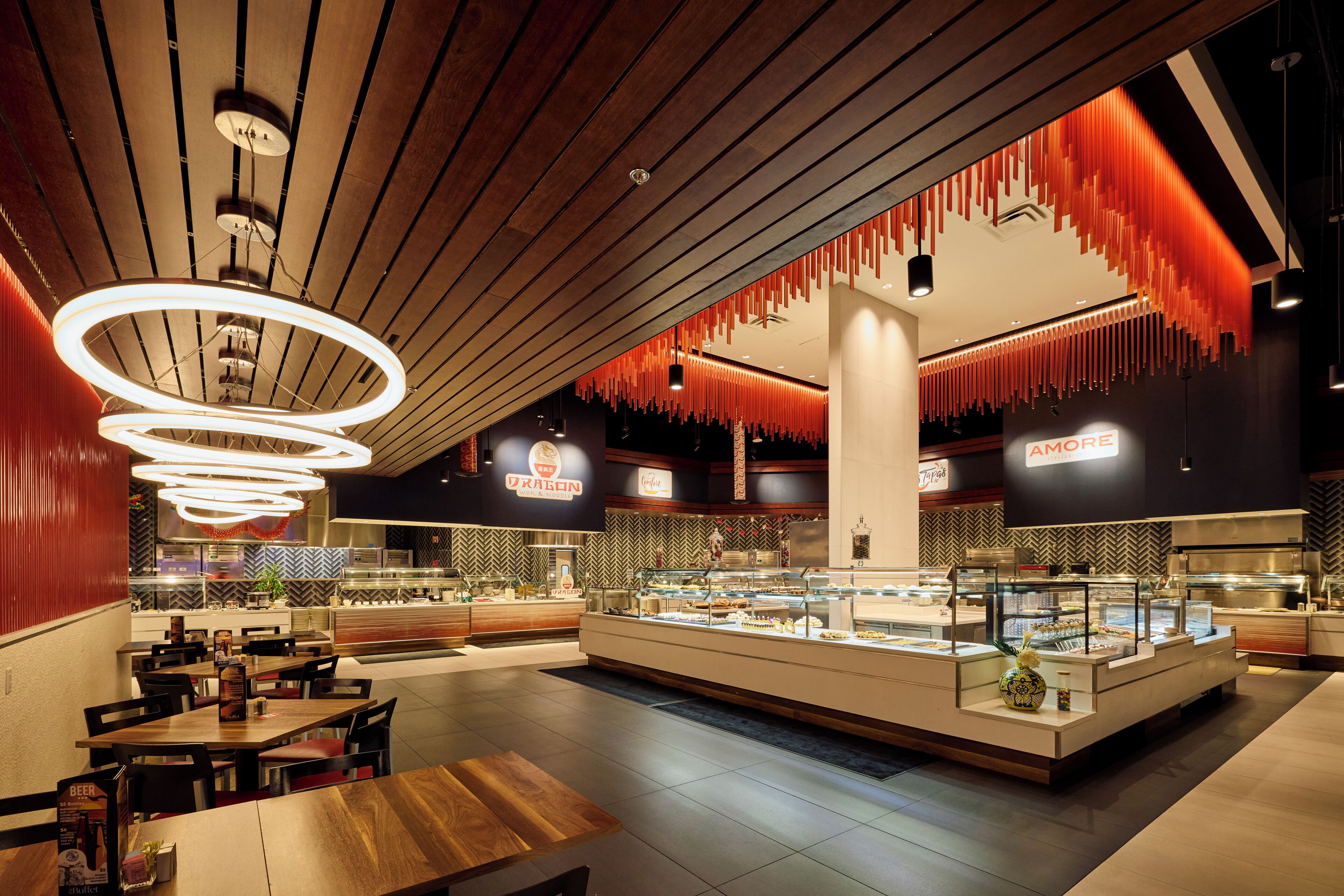 A modern restaurant interior with a central counter displaying food, wooden tables and chairs, and hanging red light fixtures.