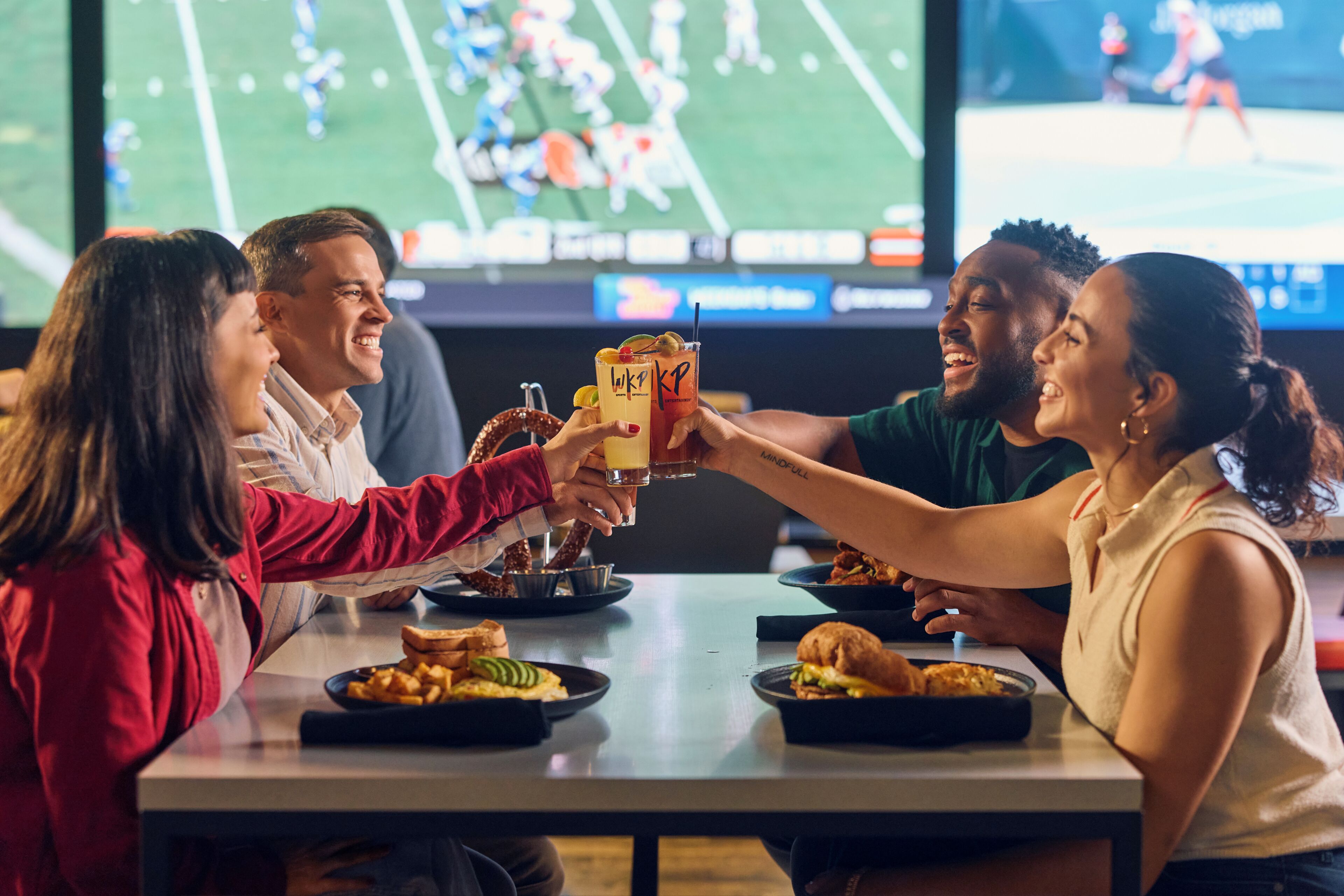 A group of people toasting with drinks at a table with food and a sports game on the TV in the background.