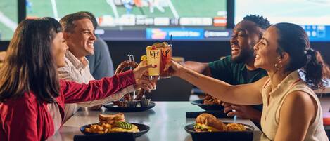 A group of people toasting with drinks at a table with food and a sports game on the TV in the background.