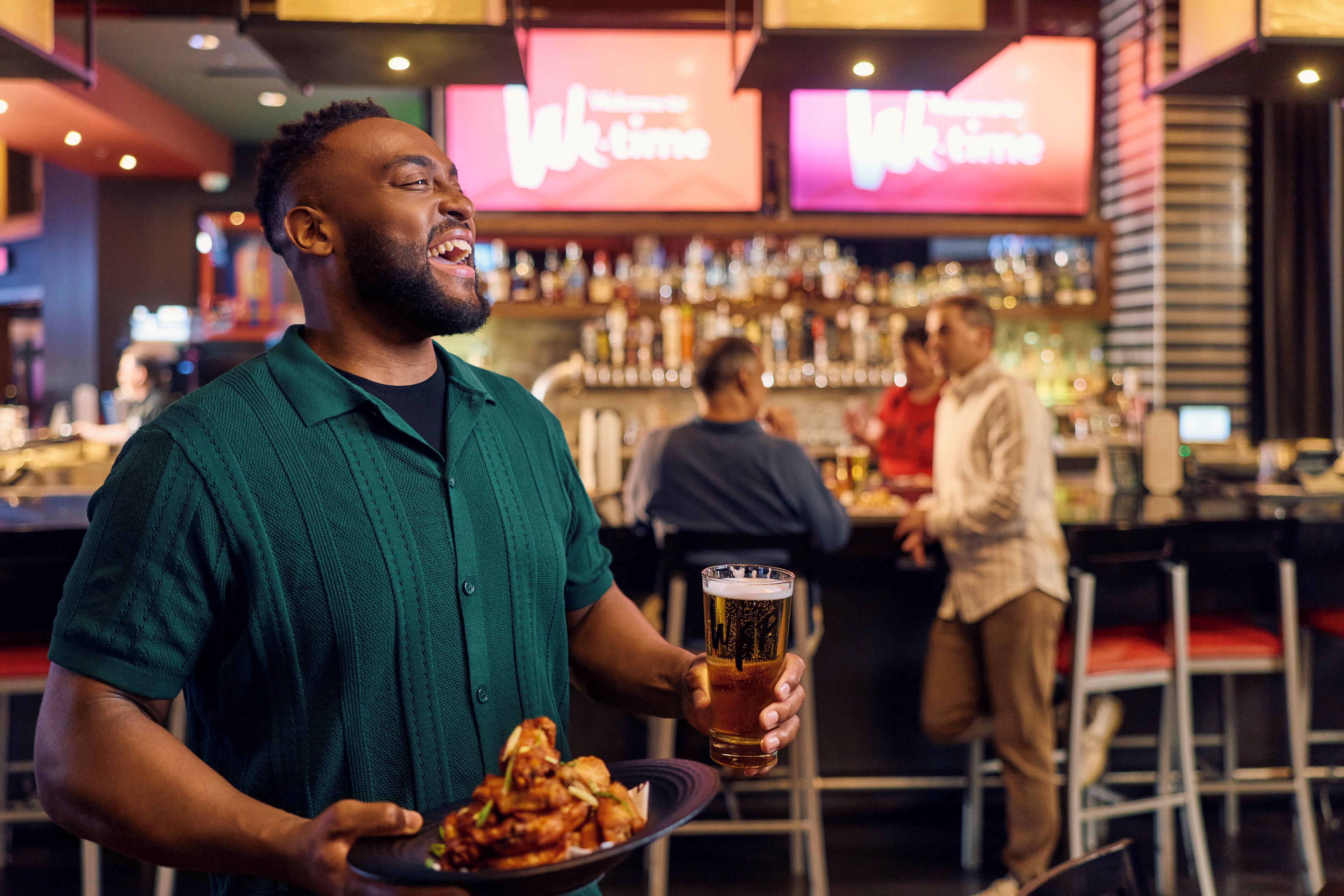 A person holding a plate of food and a glass of beer in a bar setting with shelves of bottles in the background.