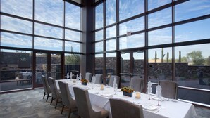 A dining area with a large table set for six, surrounded by chairs, with a view of cacti and a stone wall outside. - Wekopa Casino Resort (Fort McDowell)