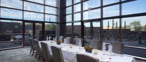 A dining area with a large table set for six, surrounded by chairs, with a view of cacti and a stone wall outside.