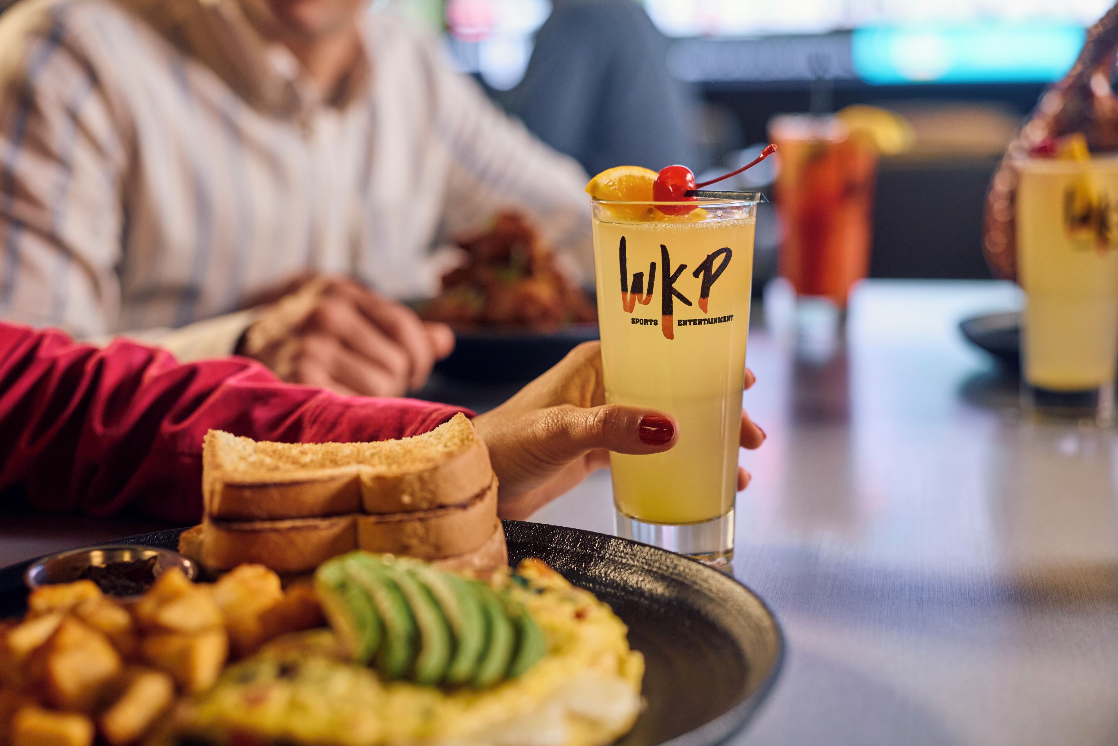 A person holding a drink with a cherry on the rim, a plate with food, and another drink in the background.