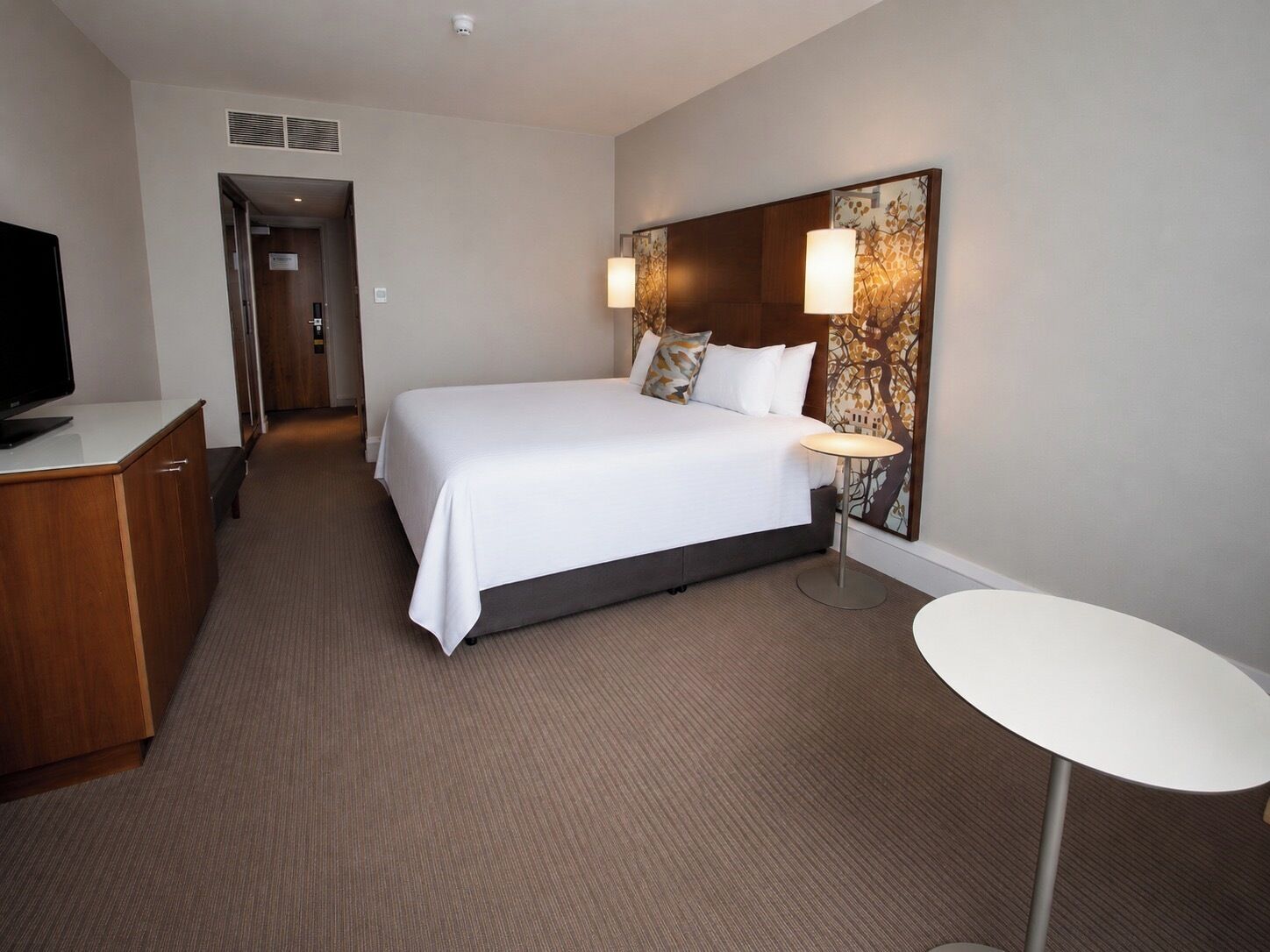 A hotel room with a bed, bedside table, lamp, and a television on a desk.