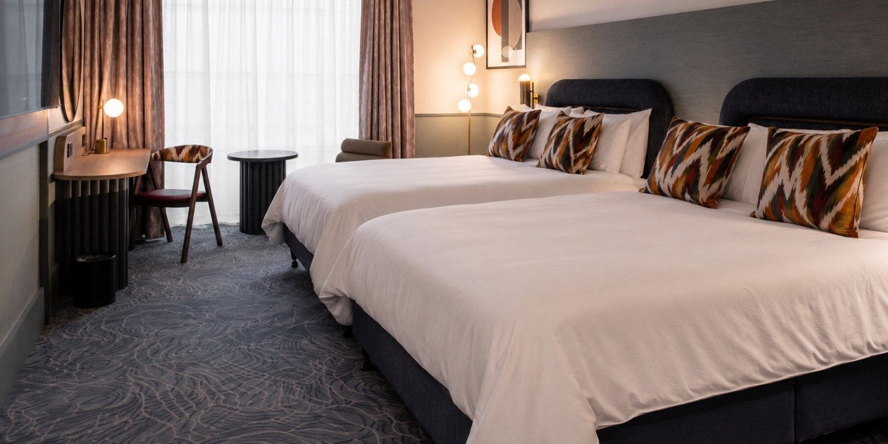 A hotel room with two beds, a desk, a chair, and a lamp.