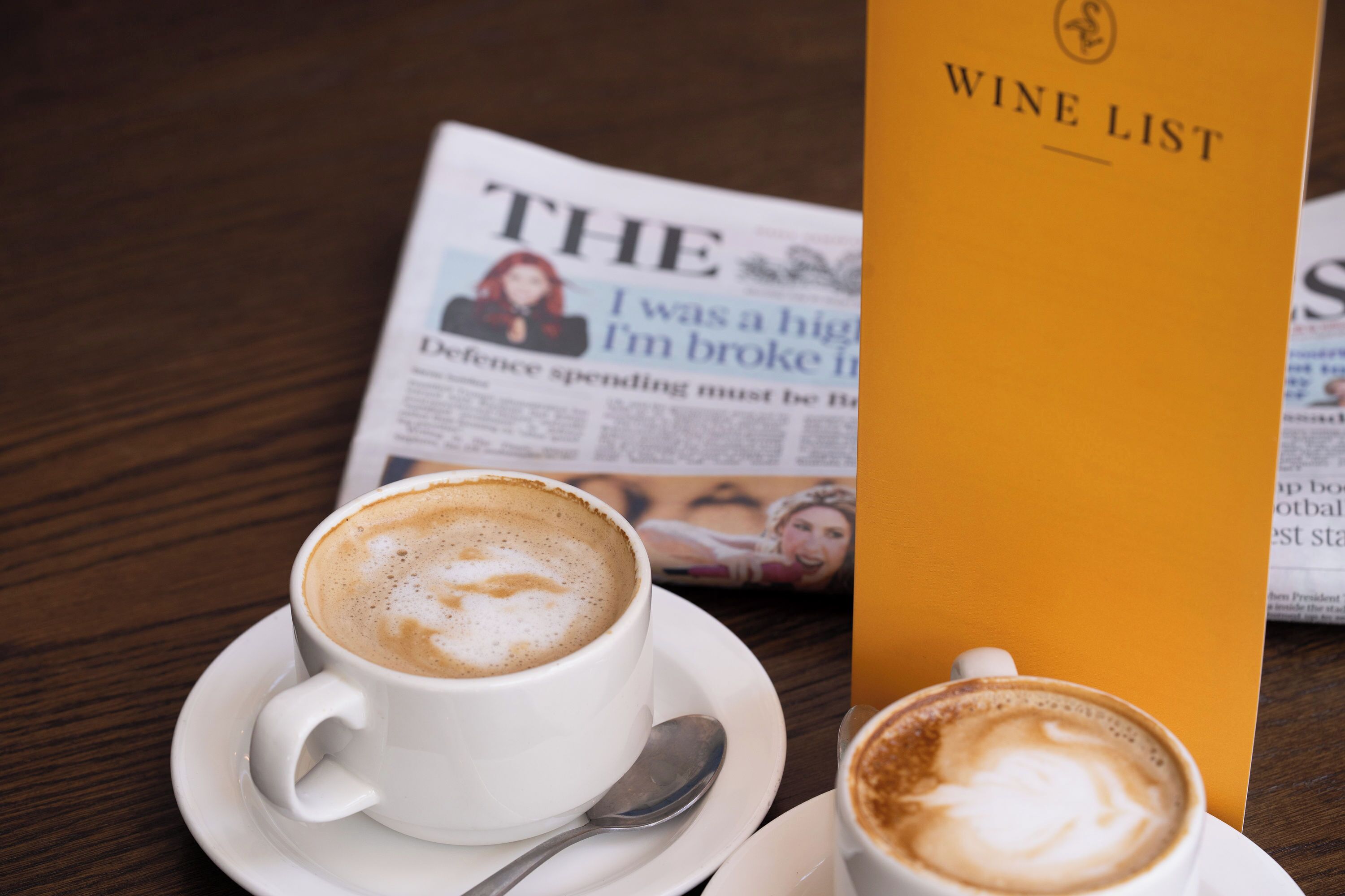 A newspaper, a wine list, and two cups of coffee on a wooden table.