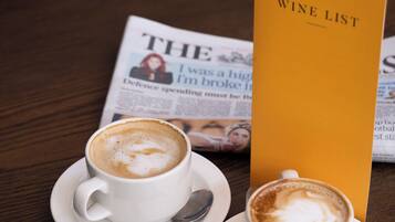 A newspaper, a wine list, and two cups of coffee on a wooden table.