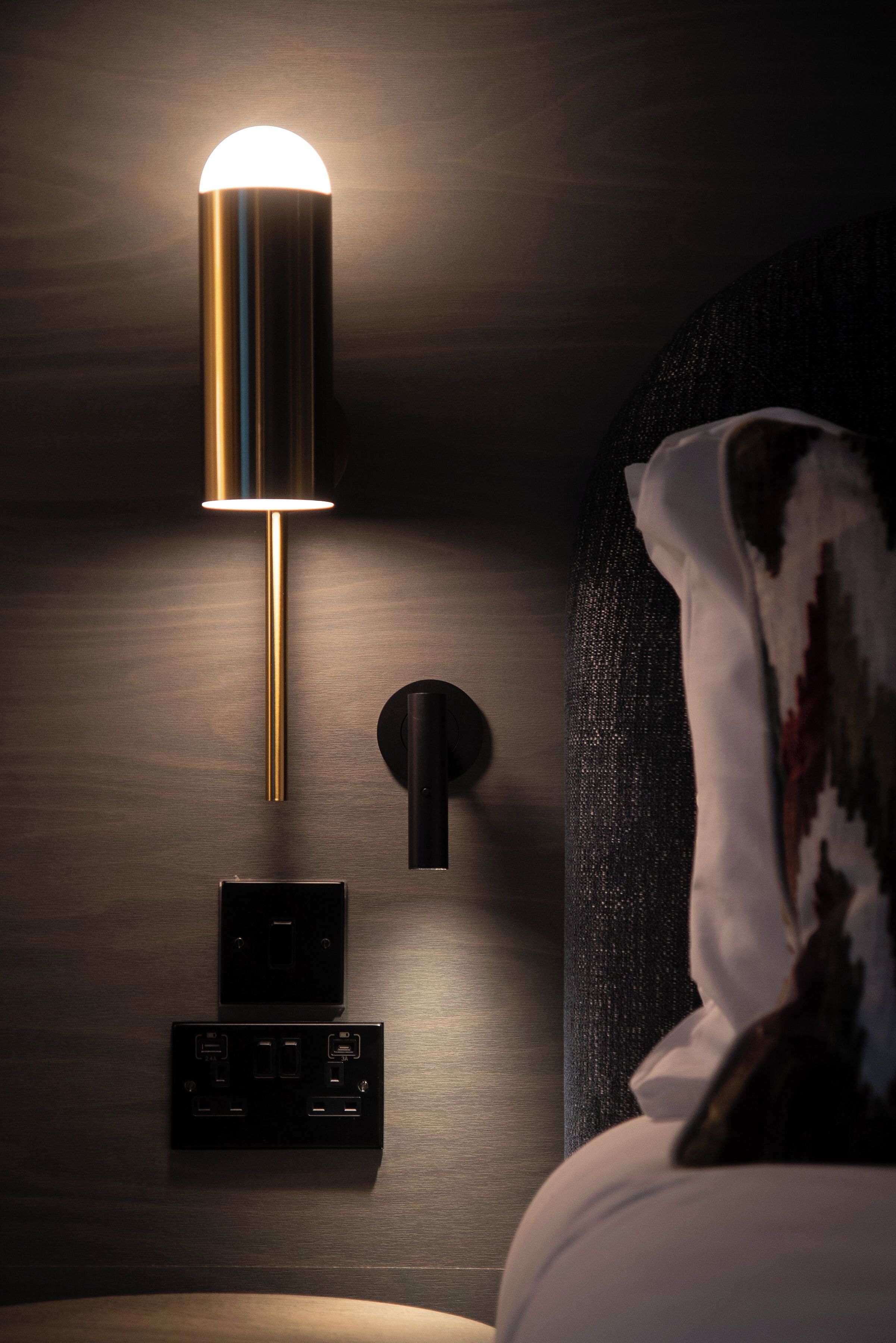 A modern wall-mounted lamp with a cylindrical shade, a power outlet, and a switch on a wooden wall.