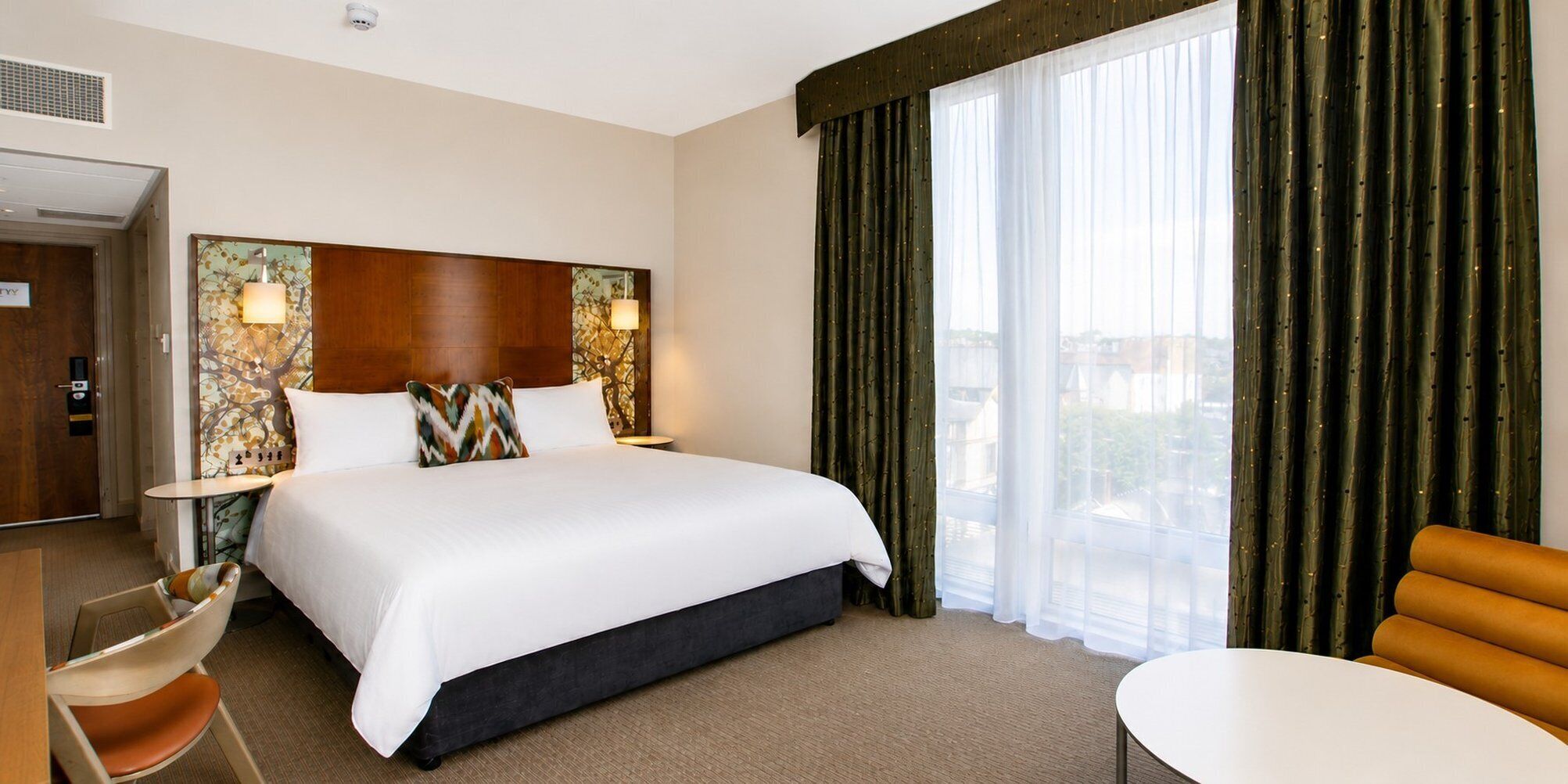 A hotel room with a large bed, a desk, a chair, and a view of the city through sheer curtains.