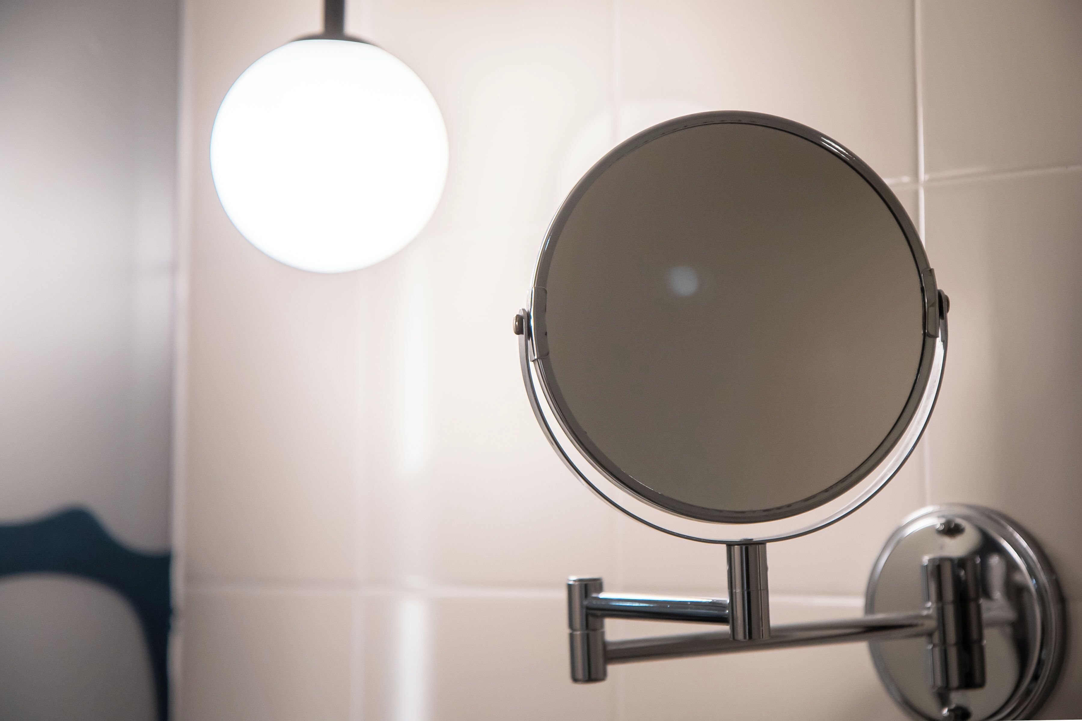 A bathroom with a round mirror mounted on a chrome stand, a round light fixture above the mirror, and a chrome faucet attached to the wall.