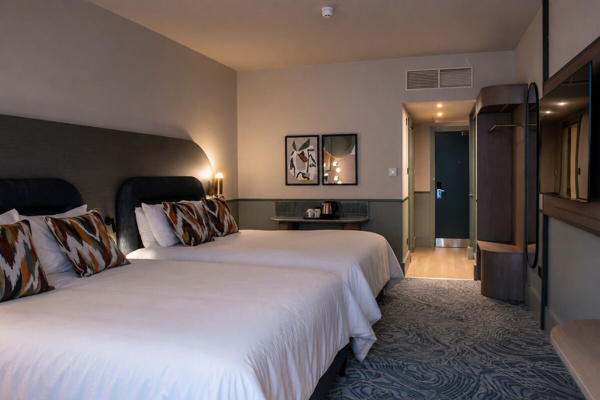 A hotel room with two beds, a bedside table, a mirror, and a door leading to a bathroom.