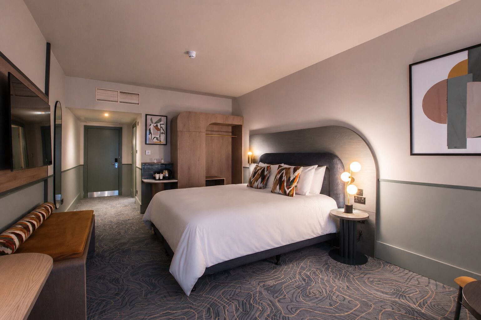 Une chambre d’hôtel avec un grand lit, une table de chevet avec une lampe, un banc et une porte donnant sur une salle de bain.