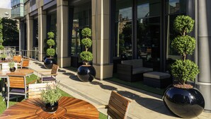 Un edificio moderno con un patio que cuenta con mesas y sillas de madera redondas, plantas en macetas y una zona de estar con banco.