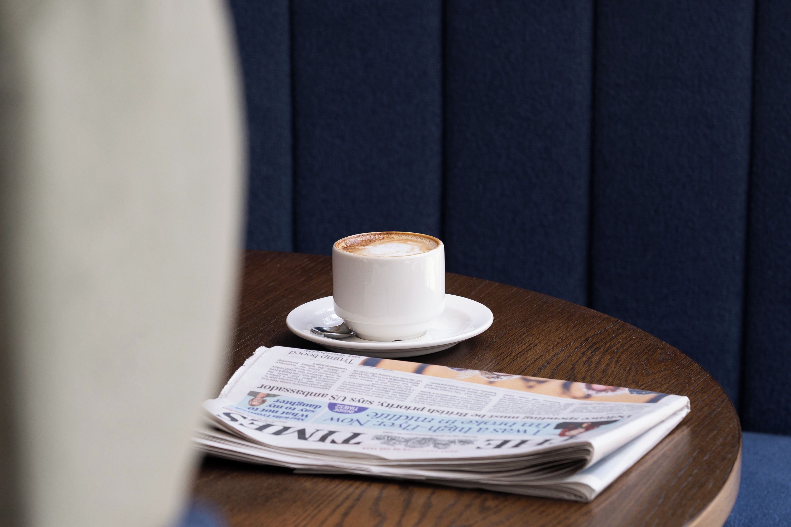 A white cup of coffee on a saucer, a spoon, and a newspaper on a wooden table.