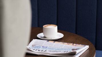 A white cup of coffee on a saucer, a spoon, and a newspaper on a wooden table.