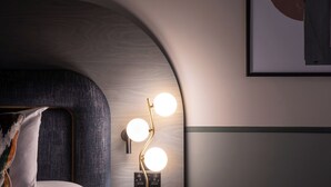Una habitación moderna con cabecera curva, una lámpara de pared con tres bombillas y una obra de arte enmarcada sobre la cama.
