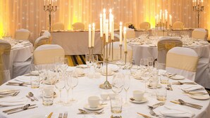 Un salón de banquetes con mesas redondas dispuestas para un evento formal, cada una con manteles blancos, velas y vajilla de porcelana fina.