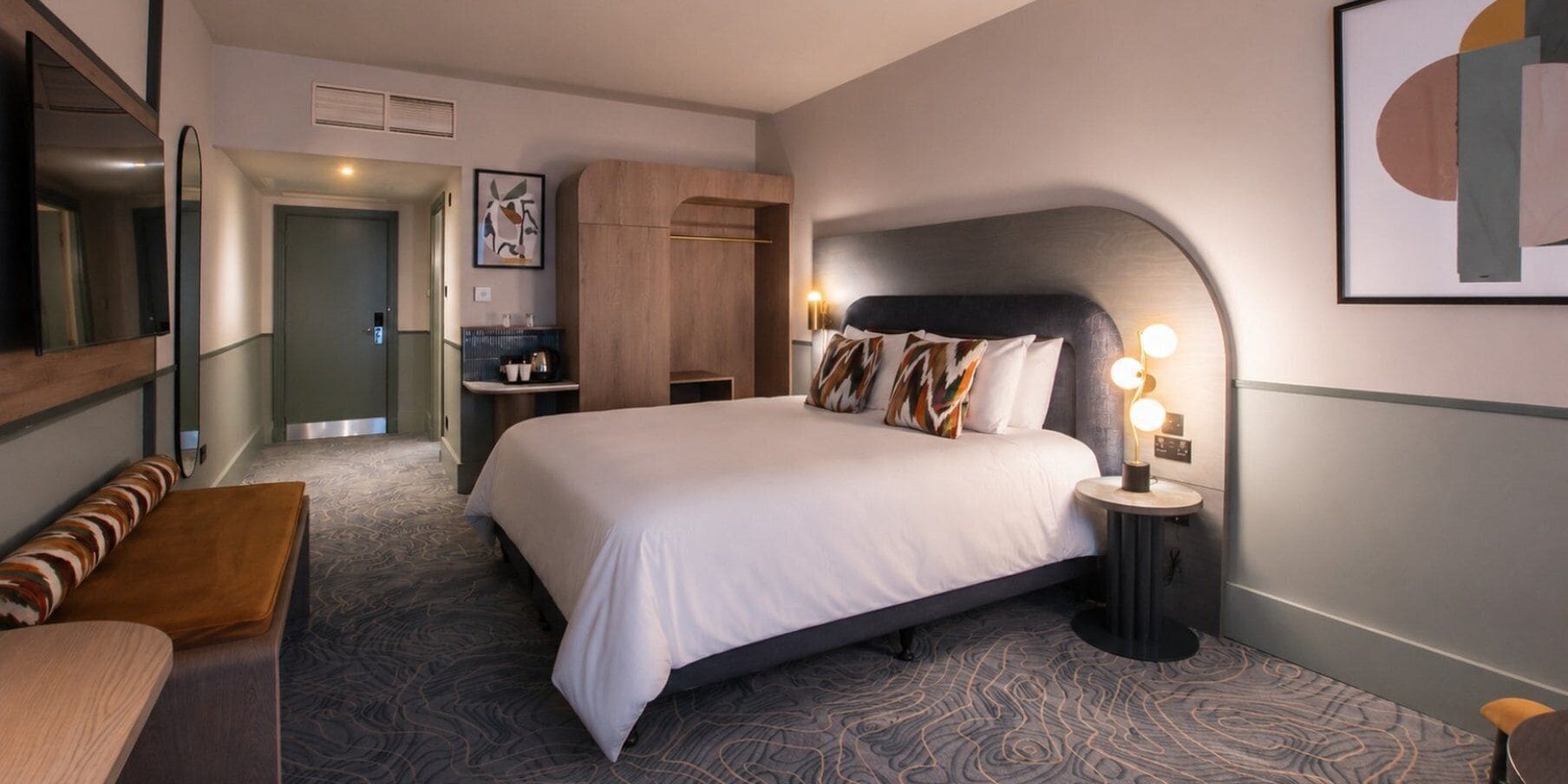 Quarto de hotel com uma cama grande, mesa de cabeceira com abajur, um banco, guarda-roupa e um espelho.