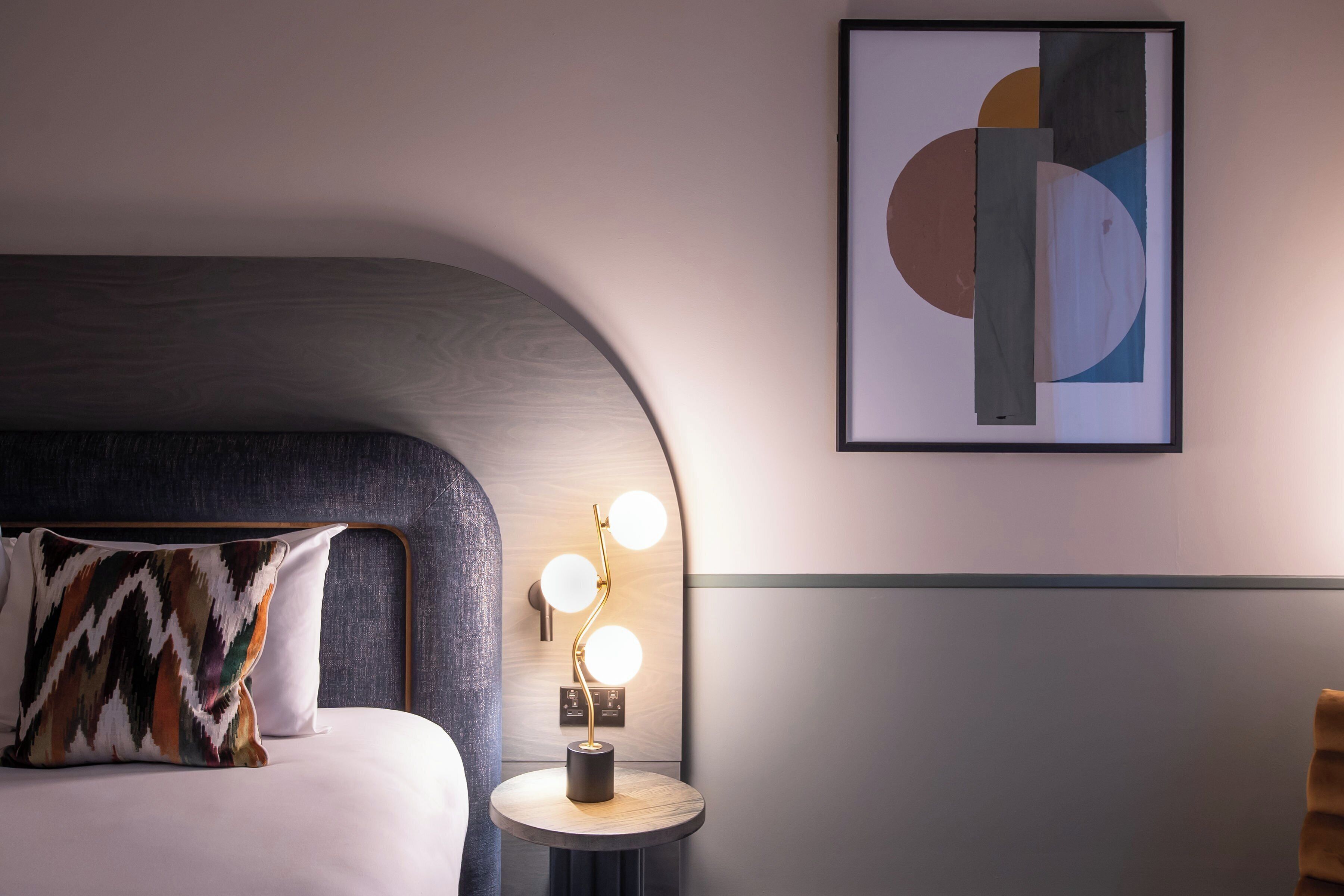 A modern hotel room with a curved headboard, a bedside table with a lamp, and a framed abstract artwork on the wall.