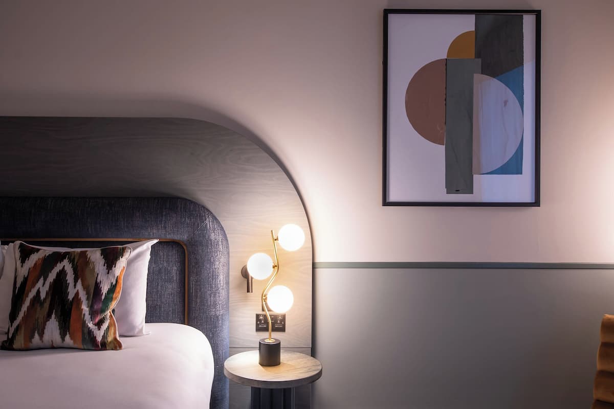 A modern hotel room with a curved headboard, a bedside table with a lamp, and a framed abstract artwork on the wall.