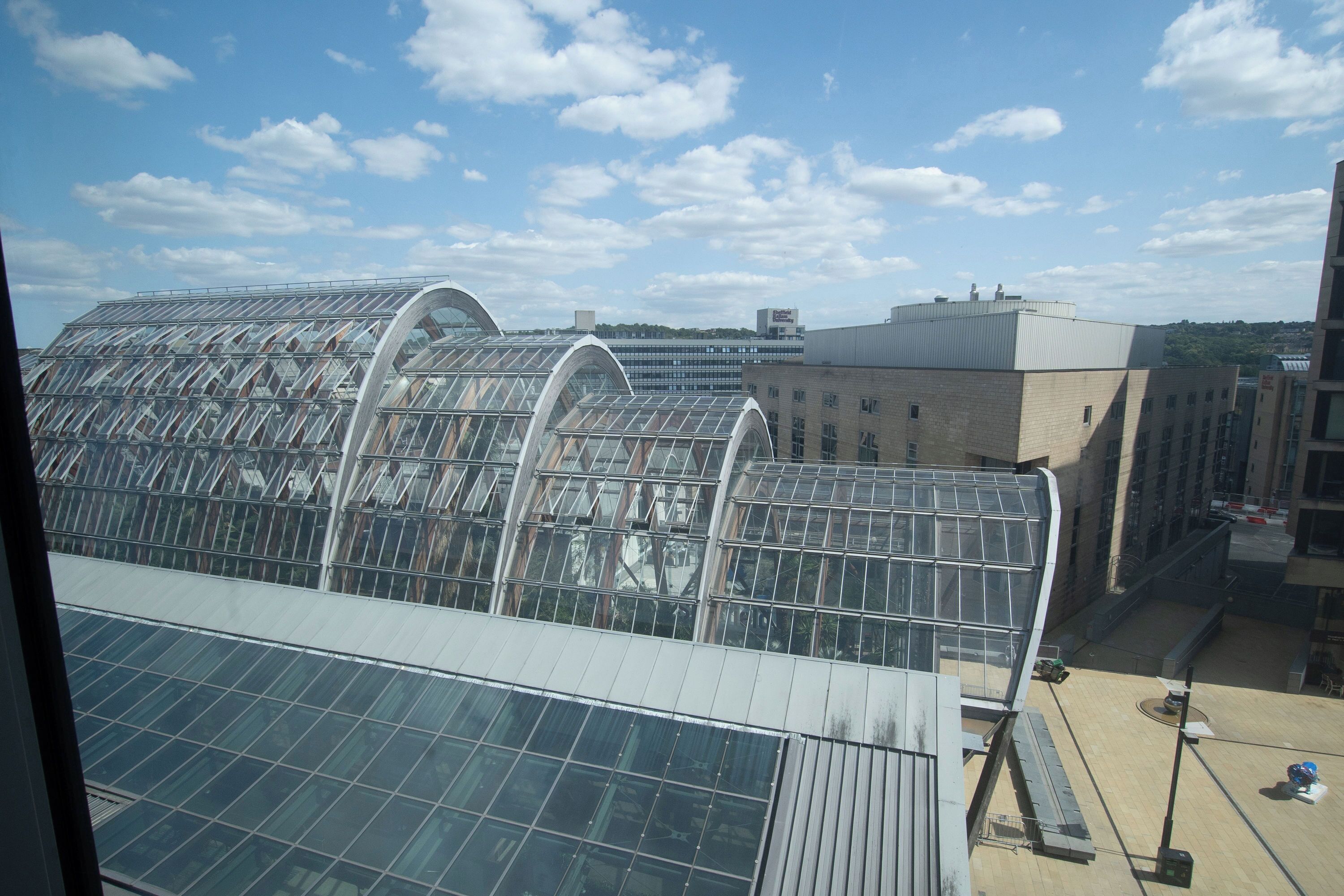A large glass greenhouse structure with a curved roof, surrounded by urban buildings.