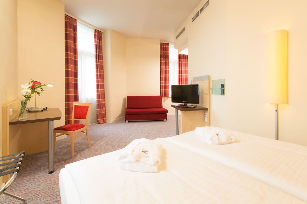 A hotel room with a bed, a desk with a chair, a television, and a red armchair.