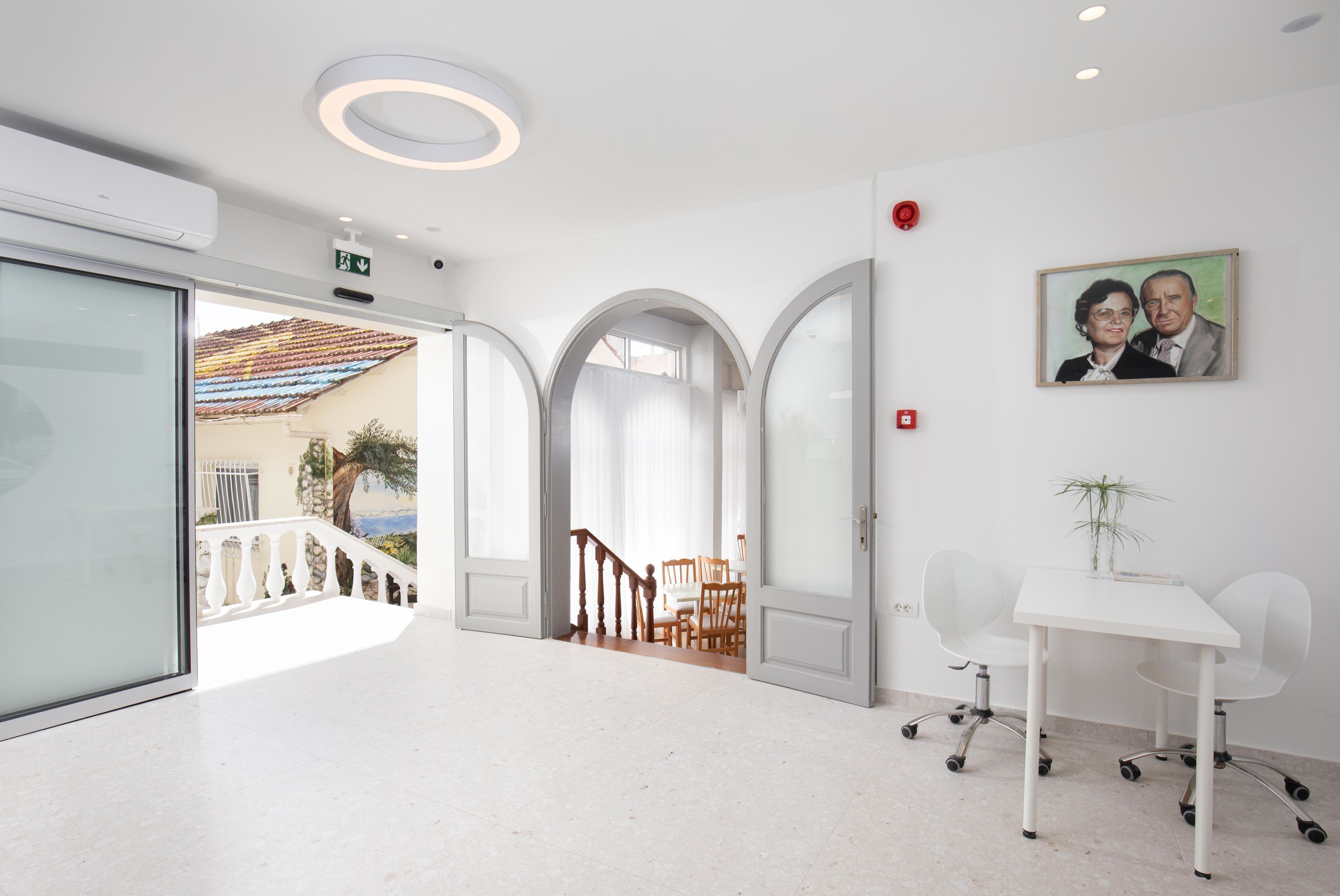 Un moderno vestíbulo de hotel con un mostrador de recepción blanco, una escalera y un área de comedor visible a través de una puerta abierta.