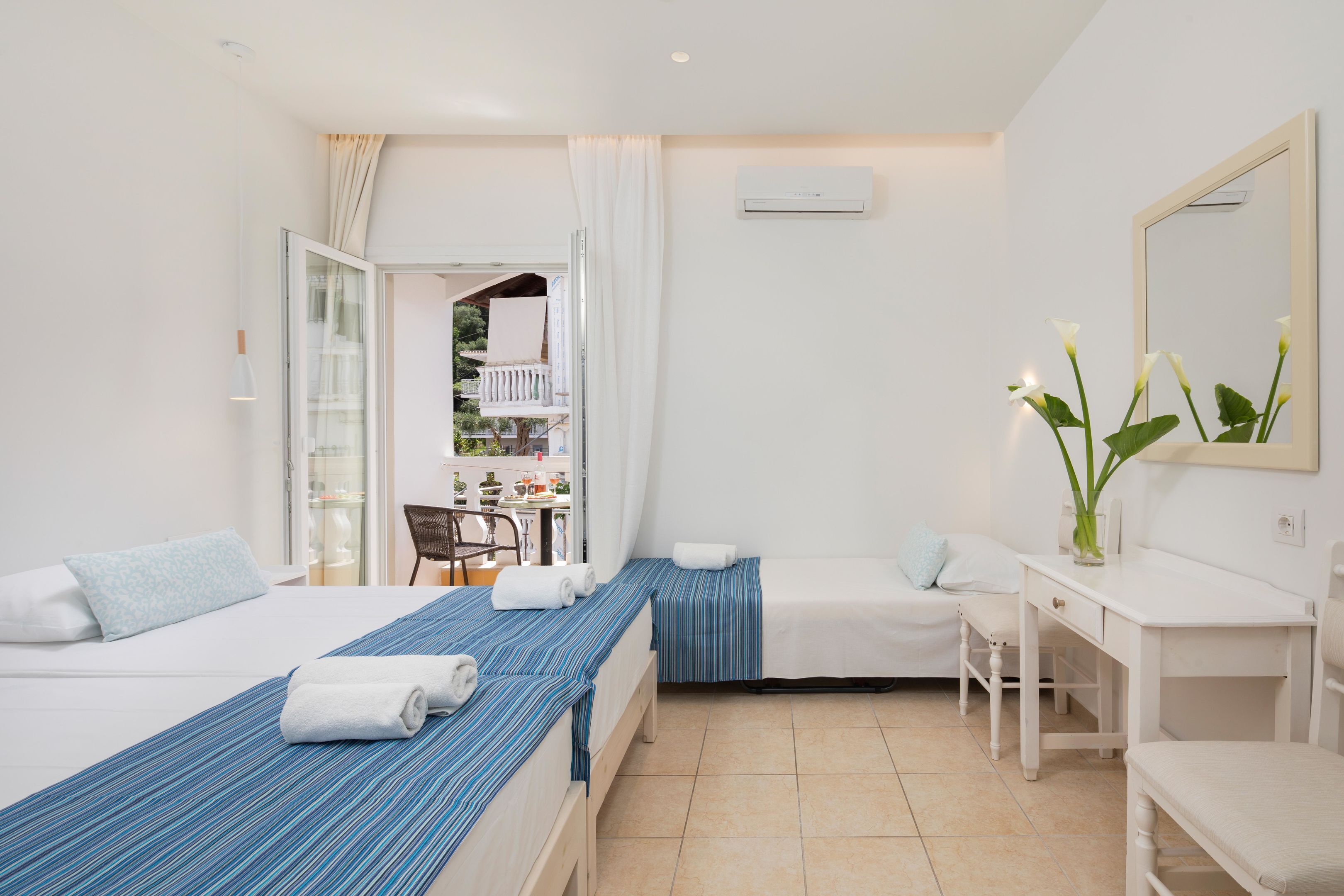 A hotel room with two beds, a desk, a chair, and a small balcony with a view of the outdoors.