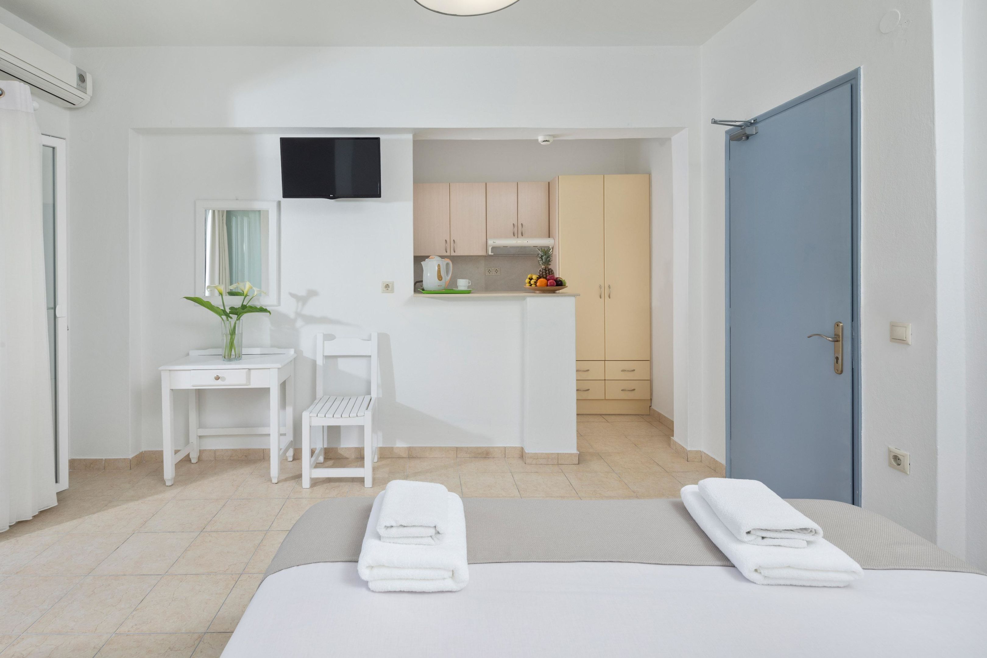 A hotel room with a bed, two white chairs, a small desk, a TV mounted on the wall, and a kitchenette area.