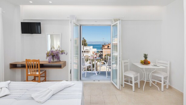 Habitación de hotel con cama, escritorio con silla, una pequeña mesa con sillas, televisor montado en la pared y balcón con mesa y sillas.