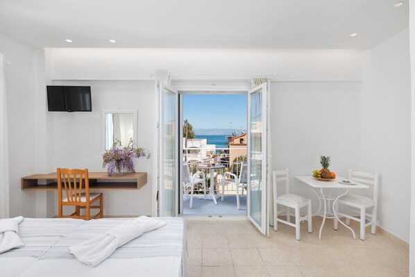 A hotel room with a bed, a desk with a chair, a small table with chairs, a TV mounted on the wall, and a balcony with a table and chairs. - Bella Vista Beach Hotel and Studios (Corfu)
