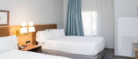 A hotel room with two beds, a nightstand with a lamp, a window with blue curtains, and a wall-mounted air conditioning unit.