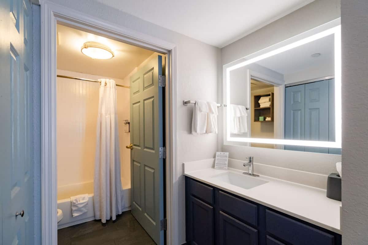 Une salle de bain avec un lavabo blanc, des meubles sombres, un grand miroir et une douche avec un rideau.