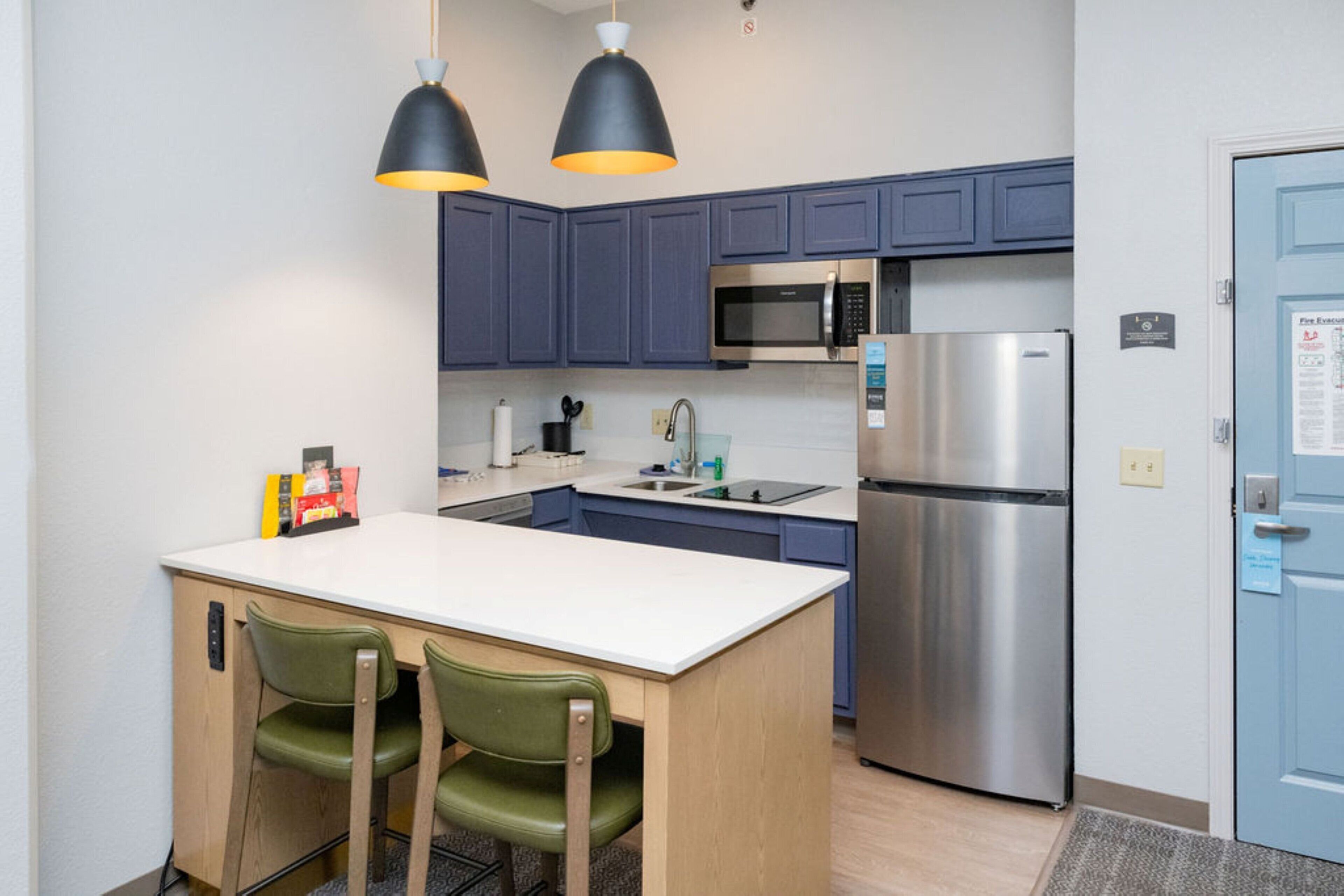 Una cocina compacta con una isla central, dos lámparas colgantes, un refrigerador de acero inoxidable y un microondas.