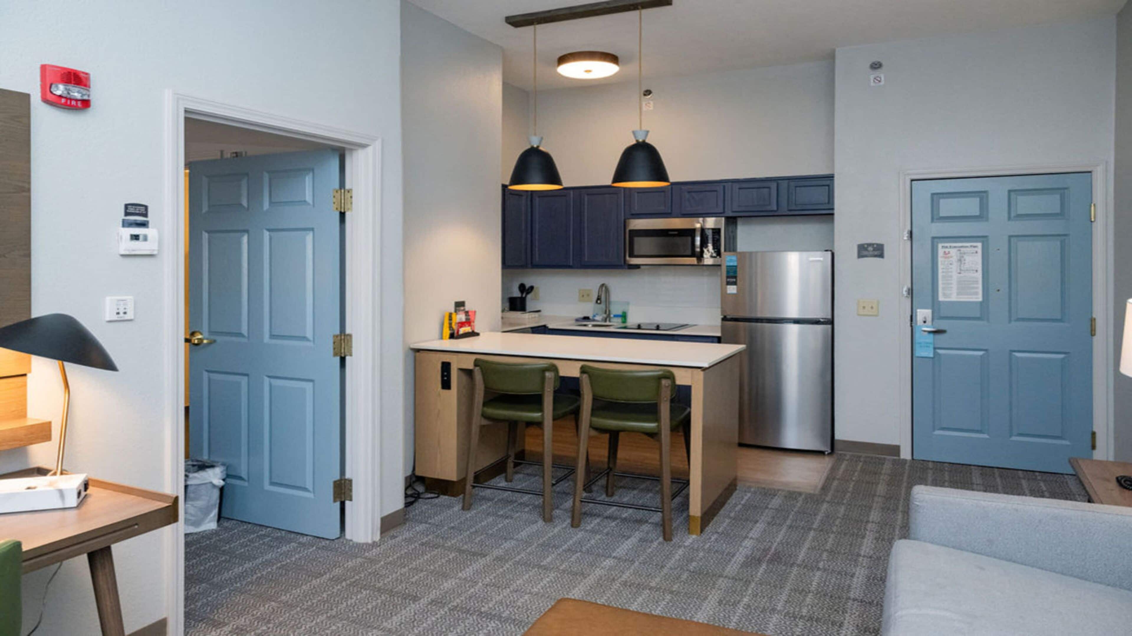 A compact kitchen with a microwave, refrigerator, and two pendant lights. There are two green chairs and a small island.